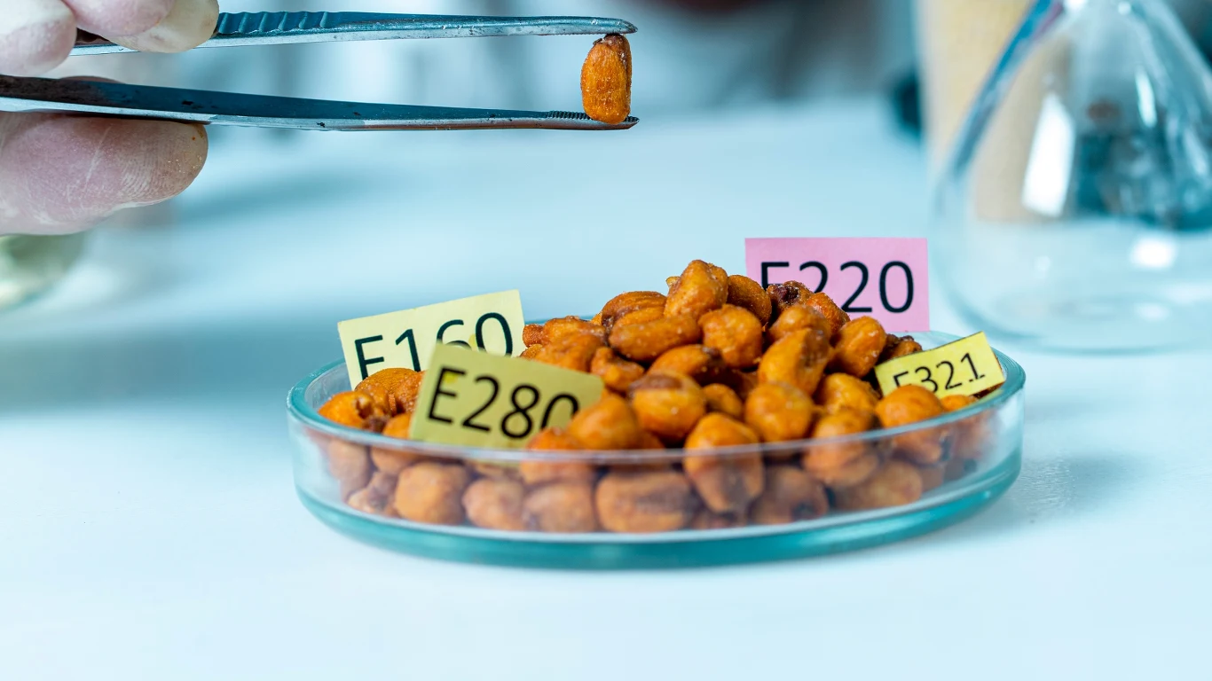 Wpływ emulgatorów na zdrowie nie jest zbyt dokładnie zbadany - zauważają naukowcy Próbka orzechów w szalce laboratoryjnej opatrzona kartonikami z oznaczeniami dodatków spożywczych E160, E280, E220 i E321, pęseta trzymająca pojedynczy orzech na pierwszym planie.