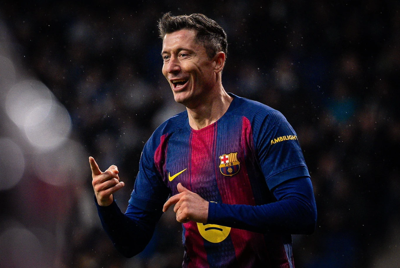 Robert Lewandowski Piłkarz w granatowo-bordowym stroju FC Barcelony podczas meczu, z uśmiechem na twarzy i uniesionymi rękami, na tle rozmazanej publiczności.