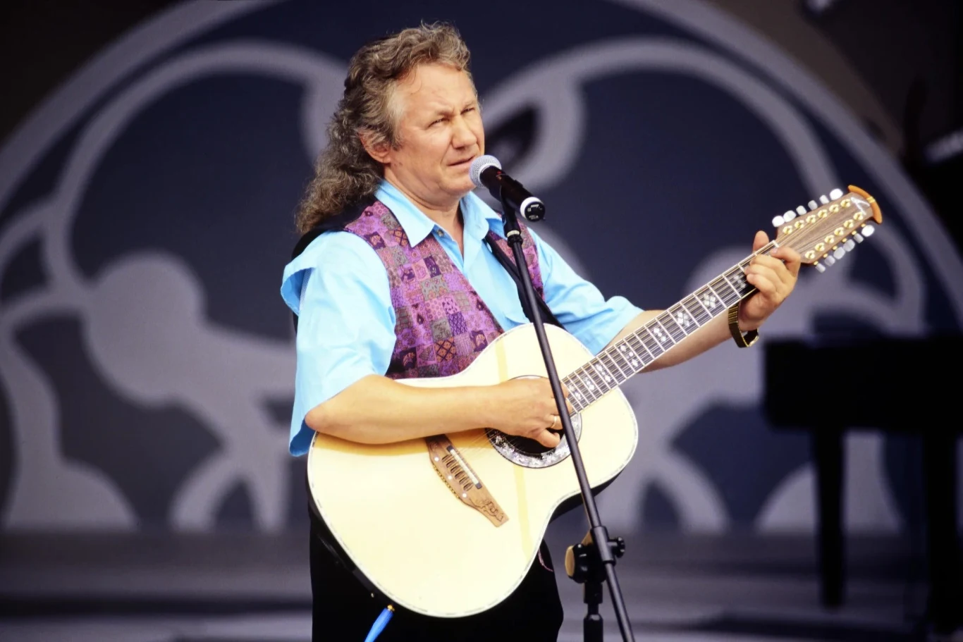 Mężczyzna w średnim wieku, ubrany w jasnoniebieską koszulę i fioletową kamizelkę, gra na dwunastostrunowej gitarze akustycznej i śpiewa do mikrofonu. W tle znajduje się rozmyte tło z abstrakcyjnym wzorem oraz fragment fortepianu.