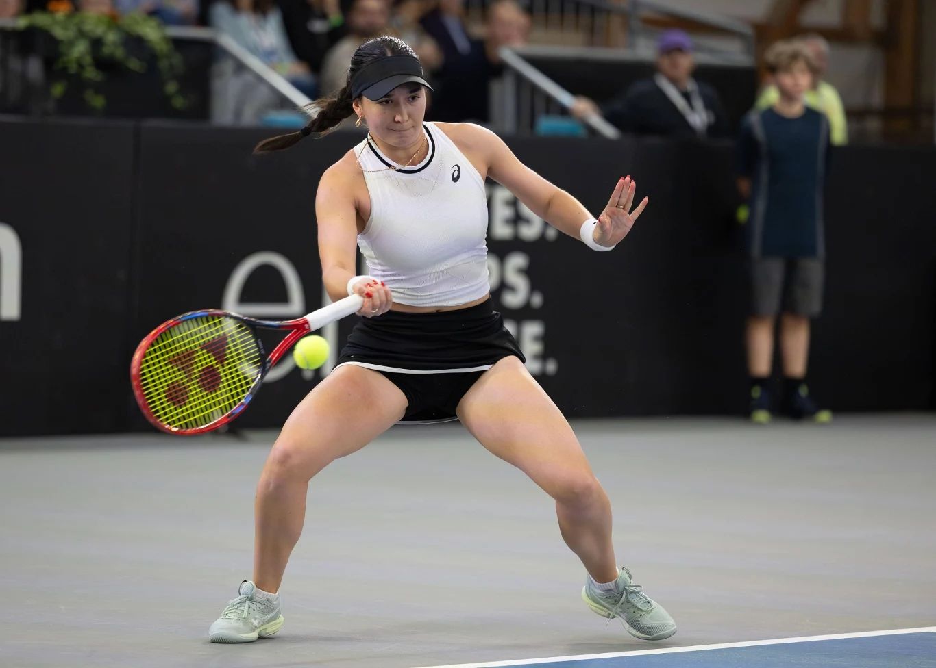 Zawodniczka tenisowa w białym stroju i czapce gra na krytym korcie, skupiona na odbiciu piłki tenisowej. W tle niewyraźni kibice oraz czarne bandy reklamowe.