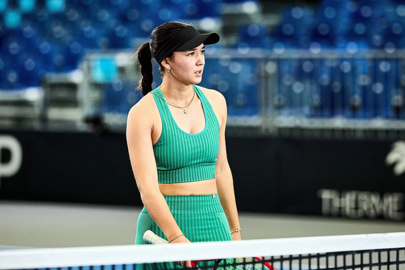 Tenisistka w zielonym stroju sportowym i czapce z daszkiem stoi przy siatce na korcie tenisowym, w tle puste niebieskie trybuny.