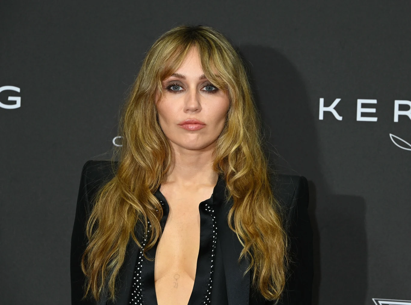 Miley Cyrus zmieniła styl ubierania, co spotkało się z wieloma pozytywnymi opiniami Kobieta z długimi, falowanymi blond włosami pozuje na tle ściany z logotypami, ubrana w ciemną marynarkę z głębokim dekoltem, wyrazisty makijaż oczu, poważny wyraz twarzy.