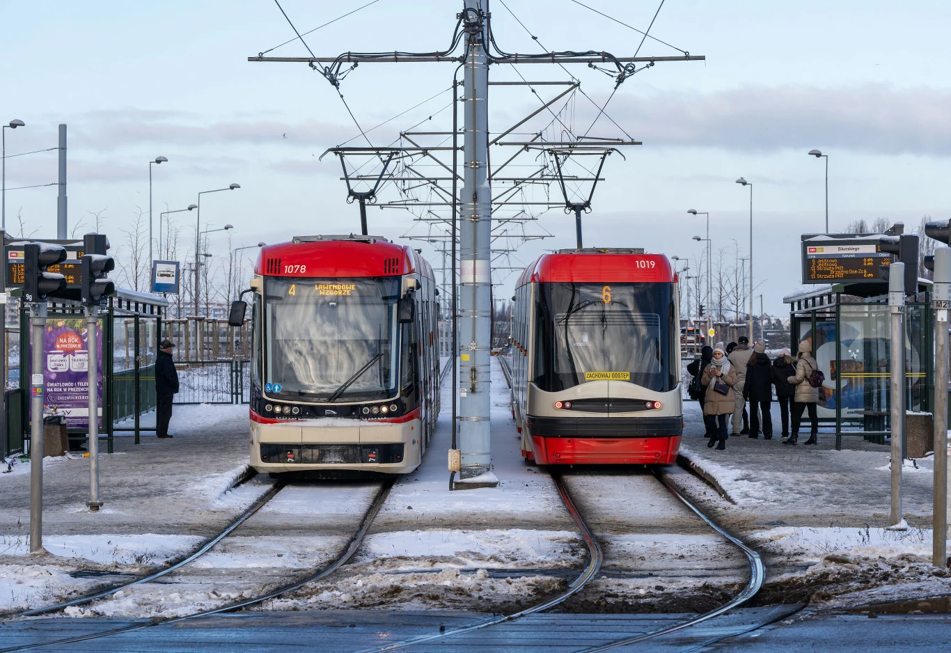 Dwa nowoczesne tramwaje stoją obok siebie na zaśnieżonym przystanku, po obu stronach widoczne są wiaty przystankowe oraz grupy oczekujących pasażerów. Zimowa atmosfera podkreślona przez leżący śnieg i ciepłe ubrania ludzi.