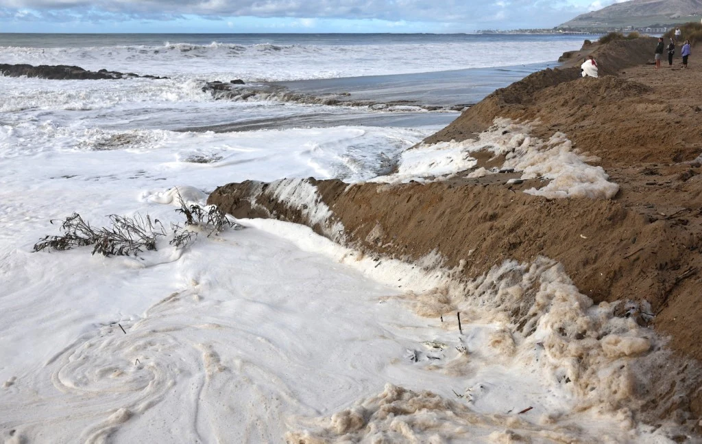Brzeg morski zebrany przez silne fale, duża ilość piany na piasku i wodzie, widoczne erozje ziemi, w tle osoby spacerujące oraz wzgórza pod zachmurzonym niebem.