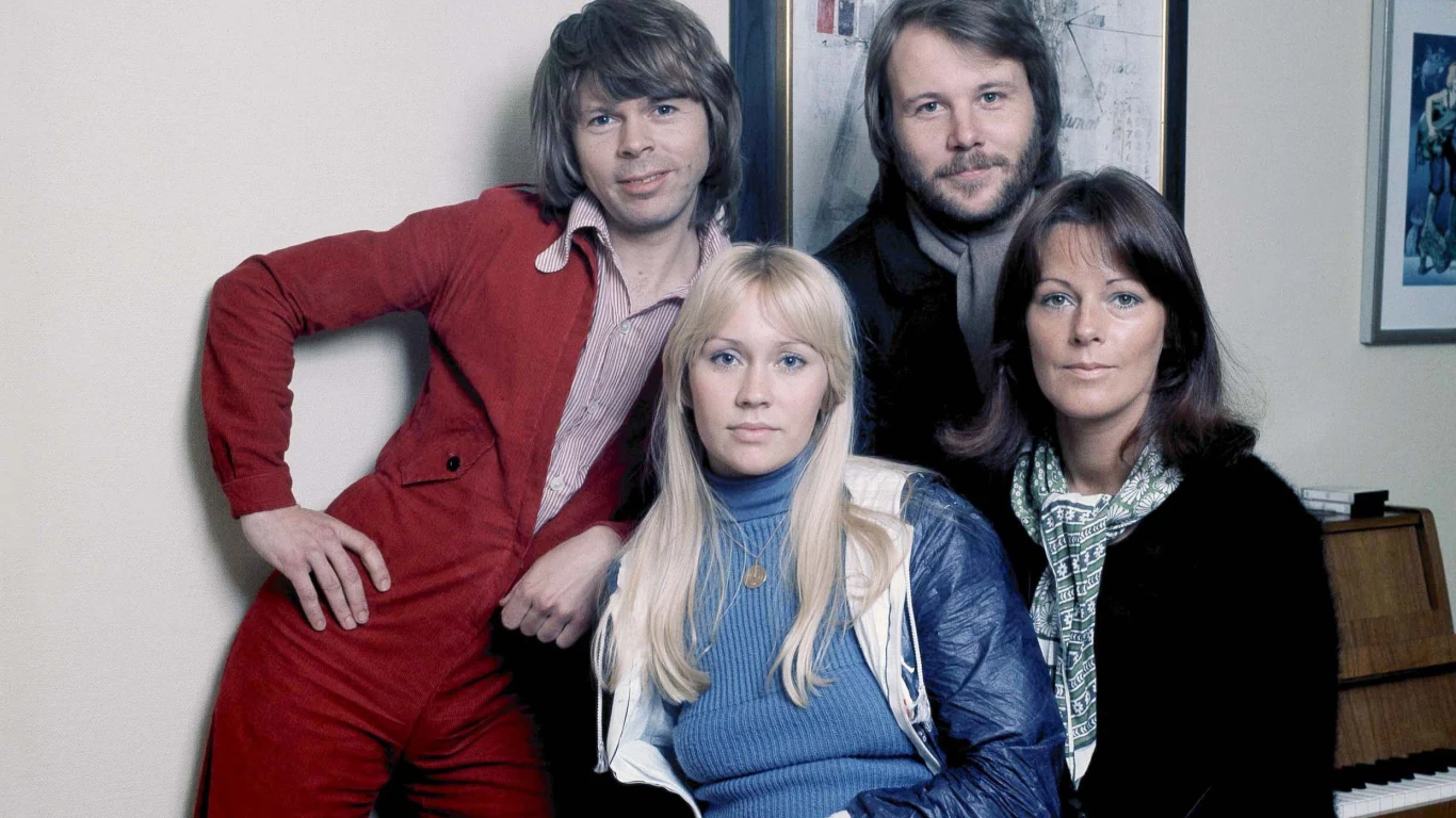 ABBA (1976), od lewej: Benny Andersson, Agnetha Faltskog, Bjorn Ulvaeus, Anni-Frid Lyngstad Cztery osoby pozujące do wspólnej fotografii w pomieszczeniu, dwie kobiety siedzą na pierwszym planie, za nimi stoją dwaj mężczyźni, w tle widoczny fragment pianina i obraz na ścianie.