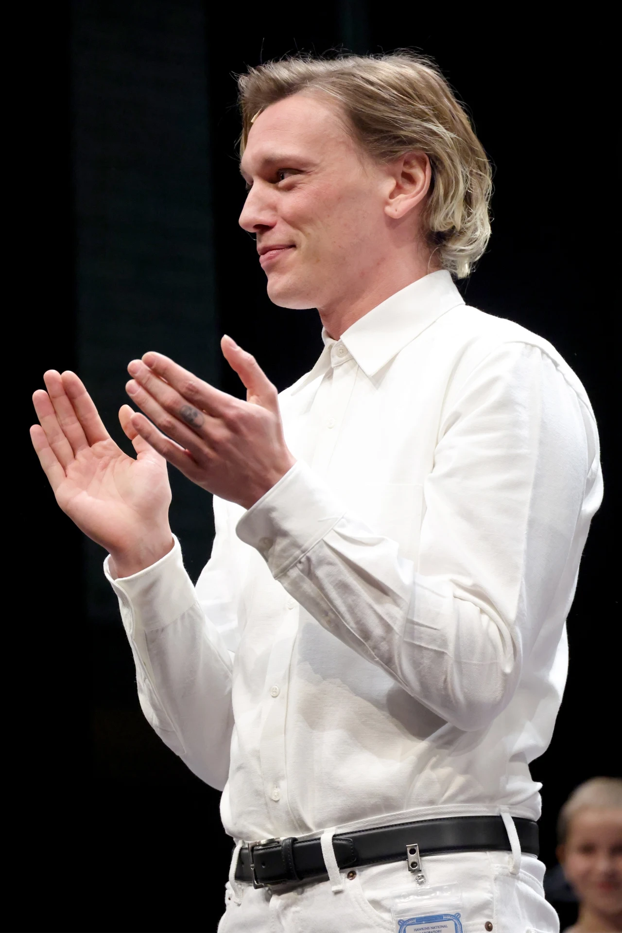 Mężczyzna w białej koszuli i białych spodniach stoi bokiem, klaszcząc w dłonie. Jasne światło pada na jego twarz i sylwetkę, a tło pozostaje ciemne i nieostre.