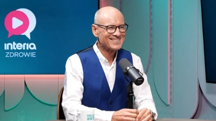 Uśmiechnięty mężczyzna w okularach i niebieskiej kamizelce siedzący przed mikrofonem w studiu, na ścianie logo Interia Zdrowie.