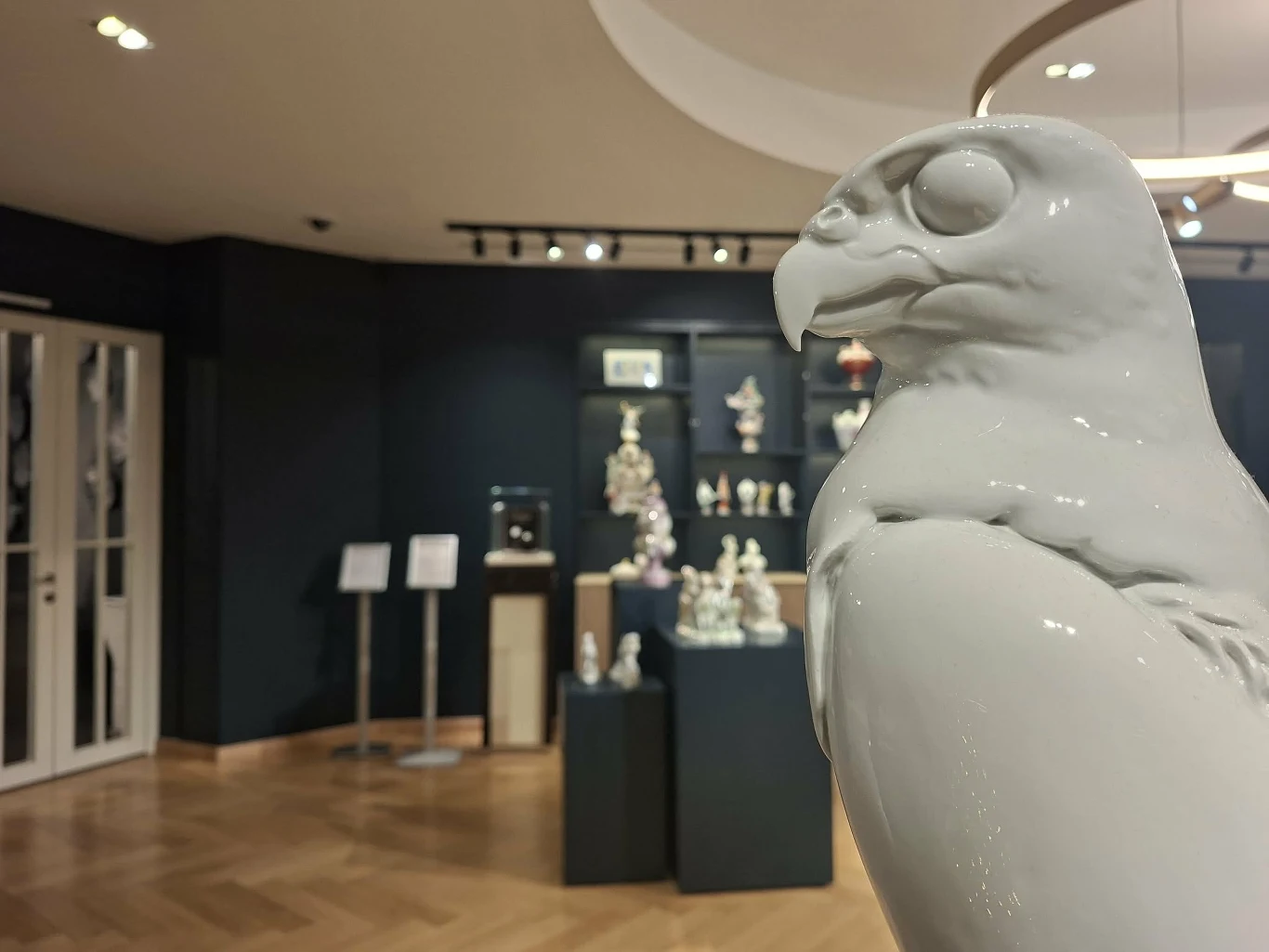 Biała porcelanowa figura ptaka, prawdopodobnie sokoła, na pierwszym planie. W tle sala muzealna z ekspozycją różnych porcelanowych rzeźb i figurek rozmieszczonych na podestach i półkach. Oświetlenie punktowe podkreśla detale oraz kompozycję wystawy.