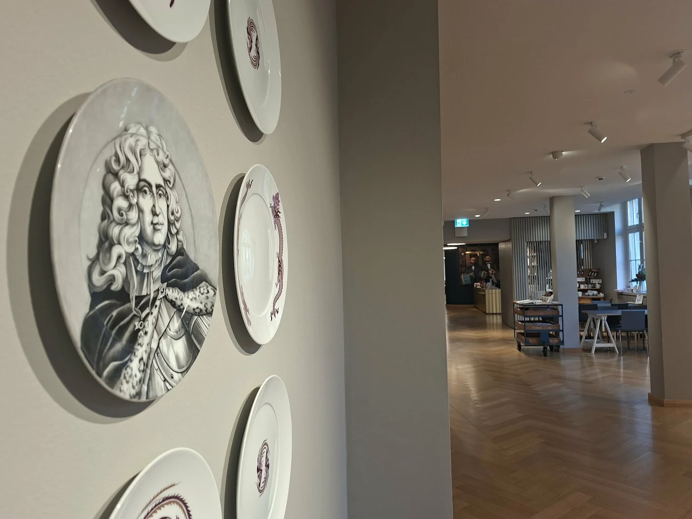 Dzięki temu, że August II Mocny interesował się wyrobami z porcelany, w Miśni powstała jej fabryka Dekoracyjne talerze na ścianie, wśród których wyróżnia się talerz z portretem historycznej postaci, z lewej strony kadru, po prawej widoczny elegancki, przestronny wnętrz budynku z drewnianą podłogą oraz meblami.