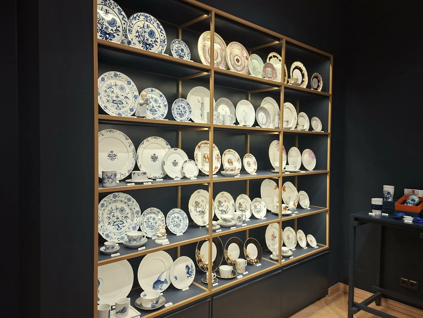 Porcelanowe talerze w Miśni Kolekcja porcelanowych talerzy i filiżanek o różnych wzorach, estetycznie ułożona na drewnianym regale w eleganckim wnętrzu, podkreślona stonowanymi ścianami i nowoczesnym oświetleniem.