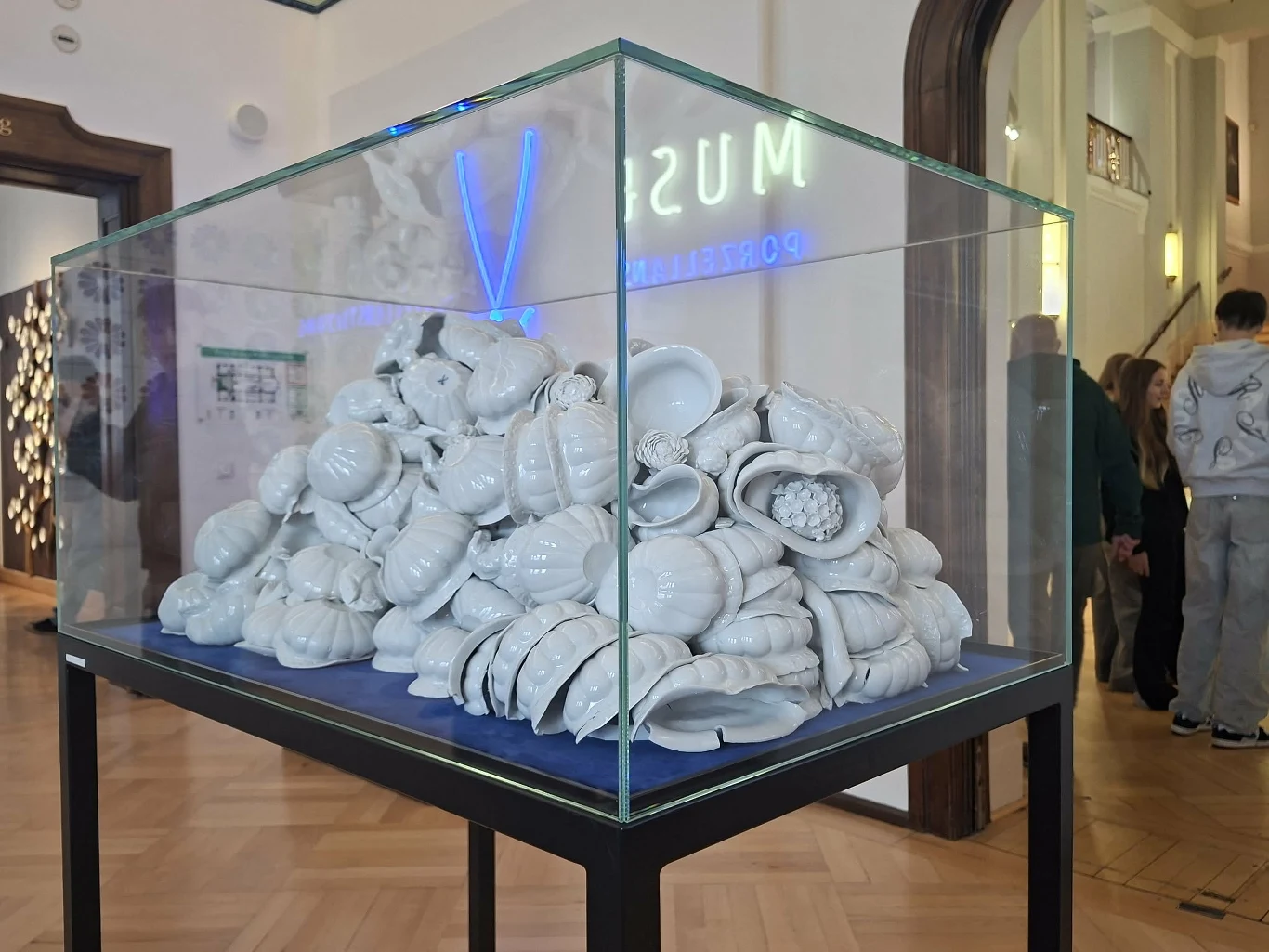 Porcelanowe rzeczy, jakie można obejrzeć w Miśni, to sztuka niekoniecznie użytkowa Stos porcelanowych masek umieszczony w szklanej gablocie muzealnej, otoczony przez minimalistyczne wnętrze z drewnianą podłogą i odwiedzających spacerujących w tle.