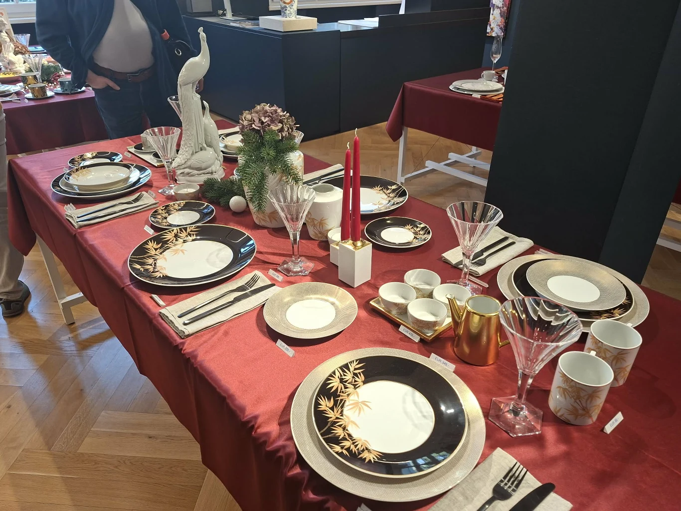 Zastawa stołowa z saskiej porcelany Elegancko nakryty stół z bordowym obrusem, porcelanową zastawą z czarnymi i złotymi zdobieniami, kieliszkami, świecznikami z czerwonymi świecami oraz dekoracjami florystycznymi, w tym figurką słonia i kompozycją z kwiatów.