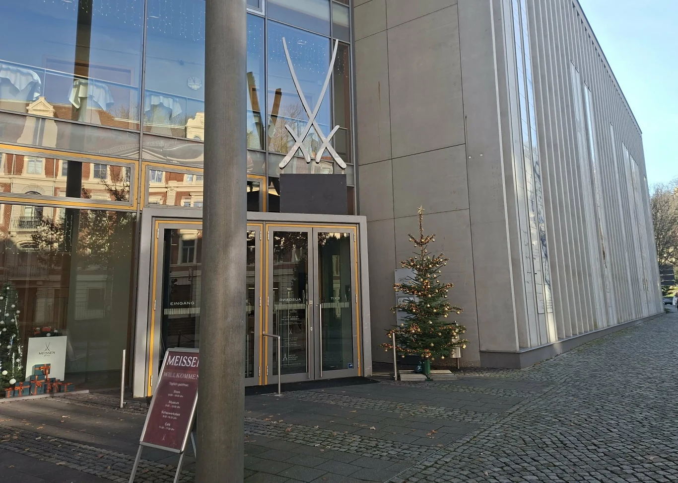 Motyw mieczy często pojawia się podczas zwiedzania Manufaktury Porcelany w Miśni Wejście do nowoczesnego budynku ozdobione dekoracją bożonarodzeniową z widoczną choinką ustawioną obok drzwi. Szklana fasada odbija pobliskie kamienice, a przy wejściu ustawiono stojak z tablicą informacyjną. Na chodniku widoczna nawierzchnia z brukowa...