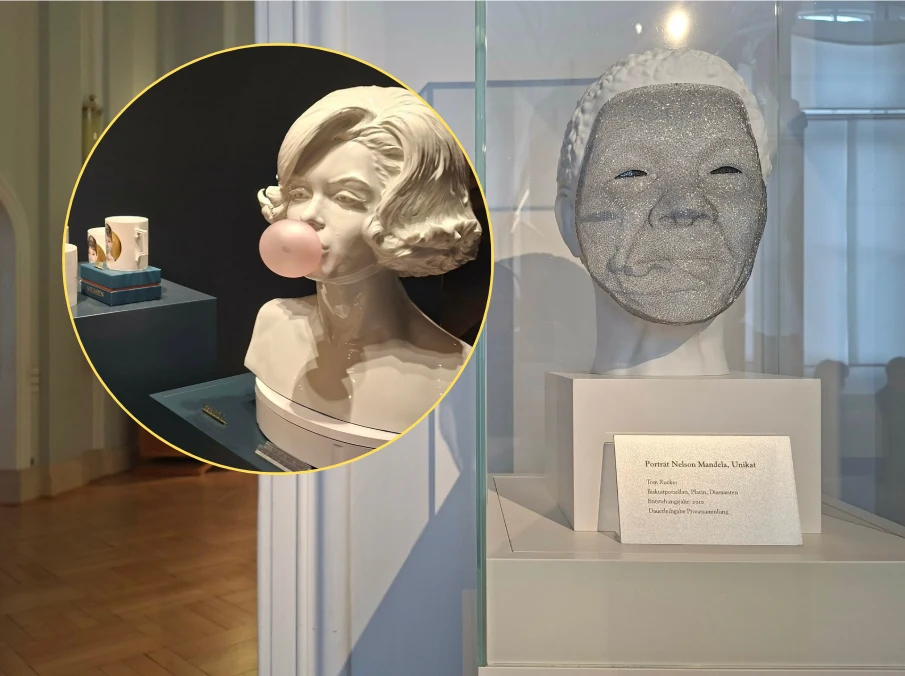 Hepburn i Mandela w wersji porcelanowej Galeria sztuki z popiersiem kobiety żującej gumę oraz szarą maską z realistycznymi rysami ludzkiej twarzy, obie rzeźby umieszczone w przeszklonych gablotach.