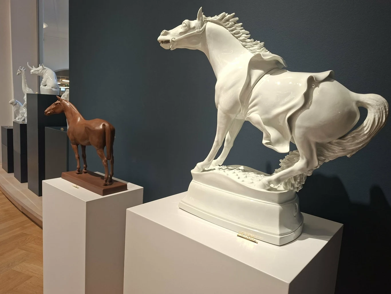 Porcelanowy koń za grube tysiące euro Kilka figur koni ustawionych na cokołach w muzeum lub galerii. Na pierwszym planie biała, dynamiczna porcelanowa rzeźba przedstawiająca galopującego konia oraz mniejsza, naturalistyczna figura brązowego konia.
