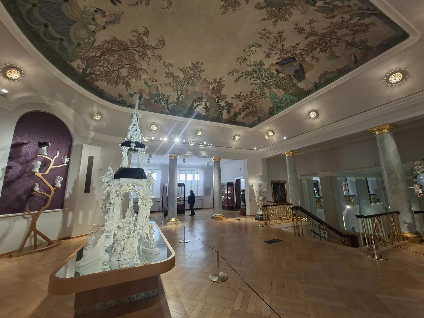 Jedna z sal muzeum w Miśni Elegancka sala muzealna z wysokim, zdobionym sufitem pokrytym malowidłem, w centralnym miejscu wystawiona biała rzeźba na podwyższeniu z lustrzanym blatem, klasyczne kolumny oraz złote zdobienia stanowią oprawę wnętrza, w tle widoczni zwiedzający.