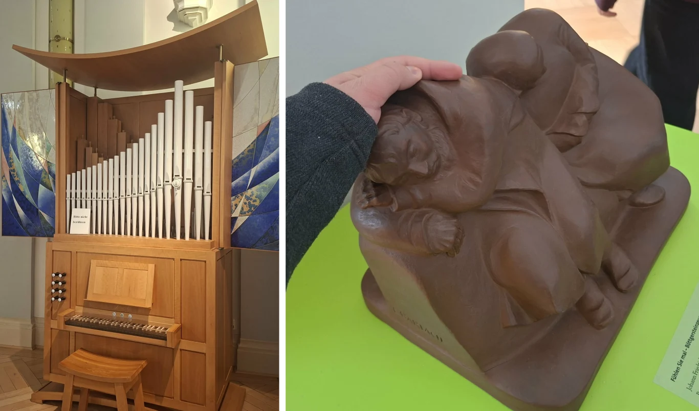 Trzeba być czujnym - z reguły eksponatów nie można dotykać, ale znalazł się wyjątek Po lewej stronie drewniane organy z licznymi pionowymi piszczałkami i malowanymi panelami w tle, po prawej ręka trzymająca rzeźbę przedstawiającą dwie osoby w geście wsparcia lub opieki, ustawioną na zielonej powierzchni.