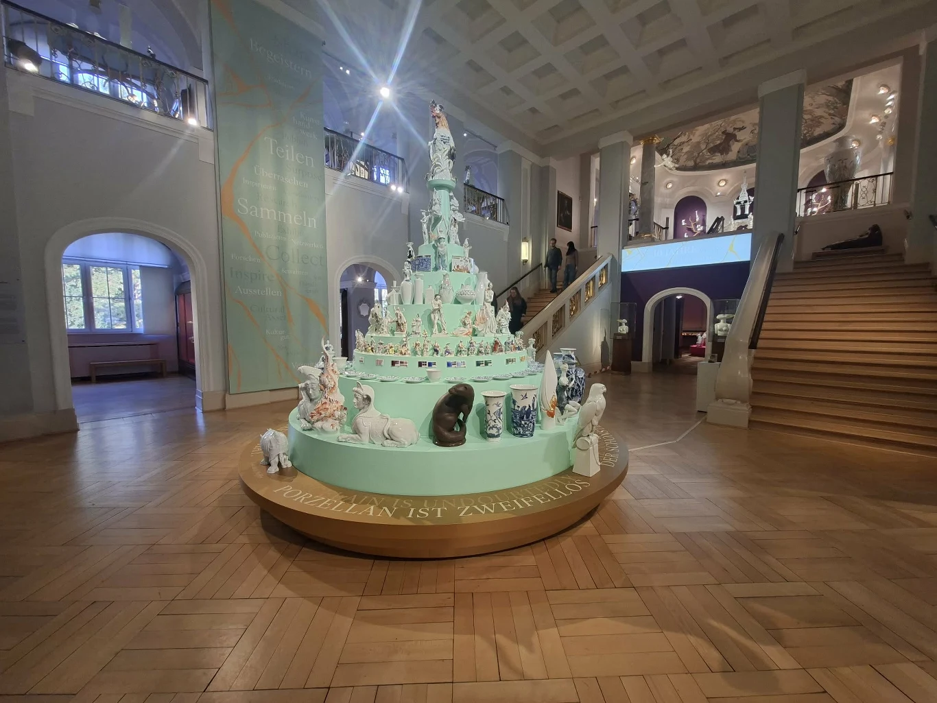 Piramidka z porcelanowymi wyrobami w Miśni Ekspozycja muzealna przedstawiająca wielopoziomową instalację z porcelanowymi figurkami zwierząt ustawioną w centralnym punkcie dużej, eleganckiej sali ze schodami. Wokół znajduje się przestronny hol z drewnianą podłogą i wysokim sufitem.