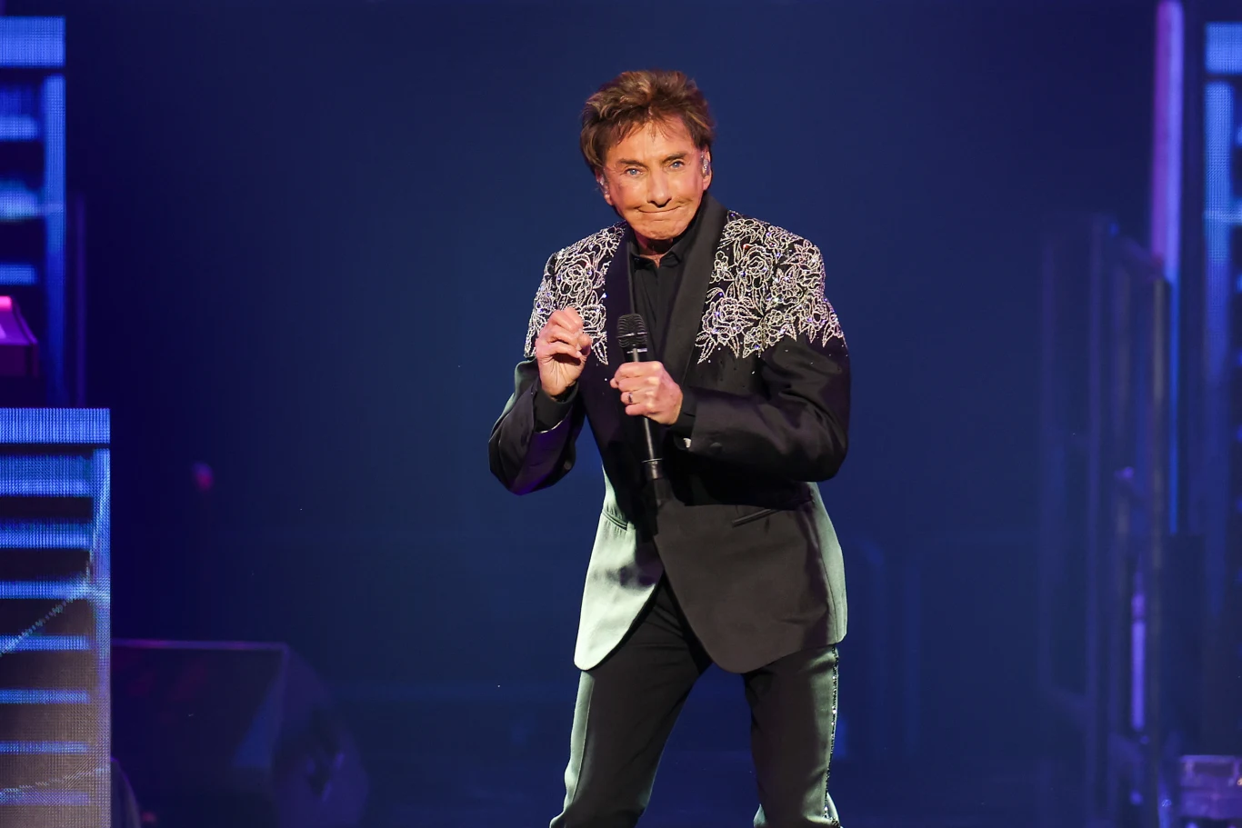 Barry Manilow przekazał najnowsze informacje o swoim stanie zdrowia Mężczyzna w eleganckim czarnym garniturze ze zdobieniami na ramionach stoi na scenie i trzyma mikrofon, tło rozświetlone światłami koncertowymi.