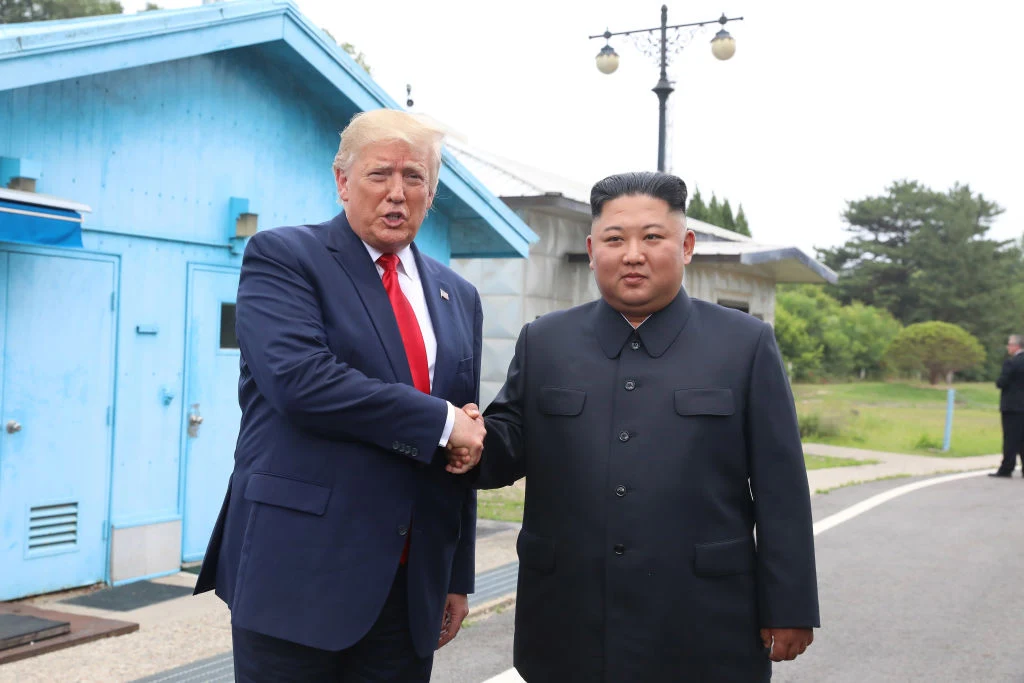 Donald Trump i Kim Jong Un podczas spotkania w 2019 roku Dwóch polityków w garniturach podaje sobie ręce na tle niebieskich budynków w strefie zdemilitaryzowanej, wokół zielona roślinność oraz w oddali widoczne są elementy infrastruktury.