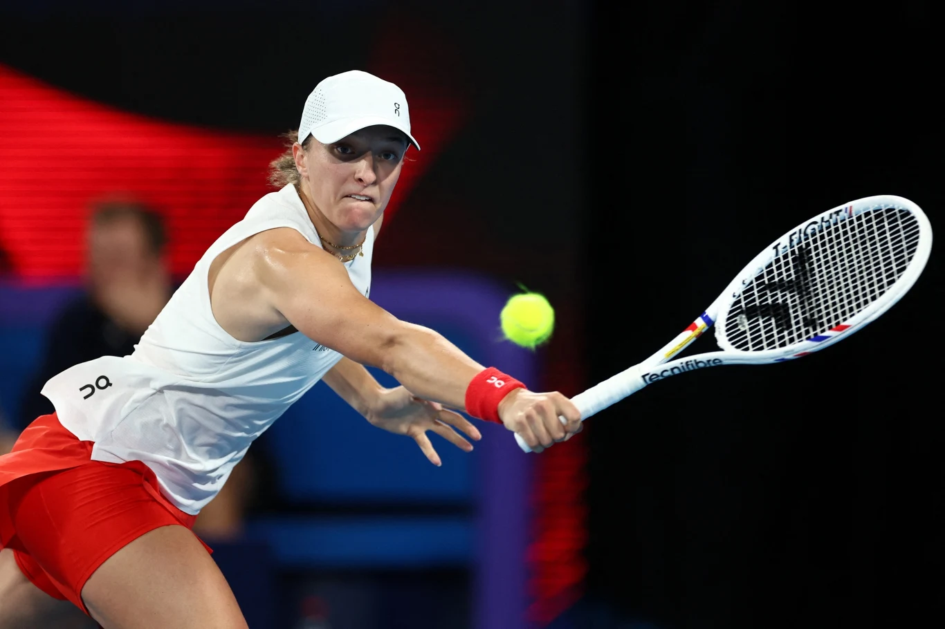 Tenisistka w białym stroju i czerwonej spódniczce odbija piłeczkę rakietą, skupiona na intensywnej grze. W tle widoczny rozmyty stadion i niebieska nawierzchnia kortu.