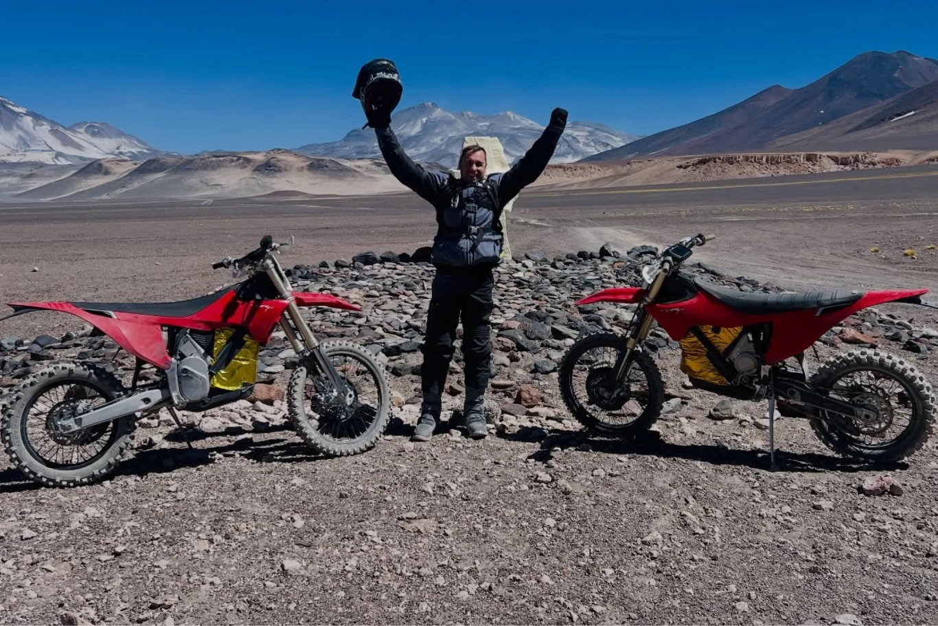 Motocyklista w ubraniu ochronnym stoi między dwoma czerwonymi motocyklami terenowymi na kamienistej pustyni, w tle rozciąga się widok na górzysty krajobraz z ośnieżonymi szczytami pod niebieskim niebem.