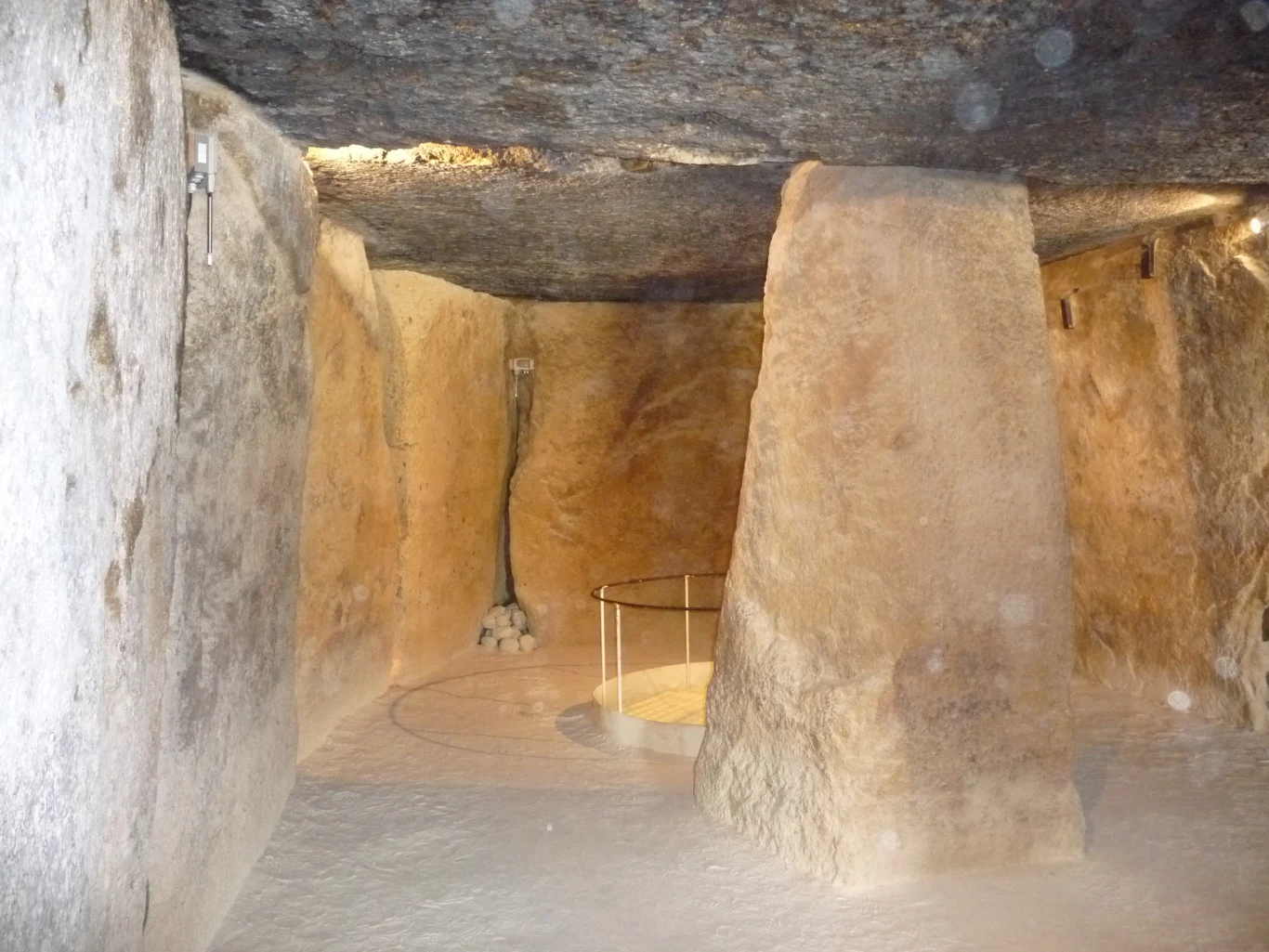 Komora wykuta w skale, otoczona masywnymi kamiennymi blokami i wspartą na centralnym filarze, z okrągłą barierką ochronną w głębi, oświetlona dyskretnie światłem wydobywającym się ze szczeliny w stropie.