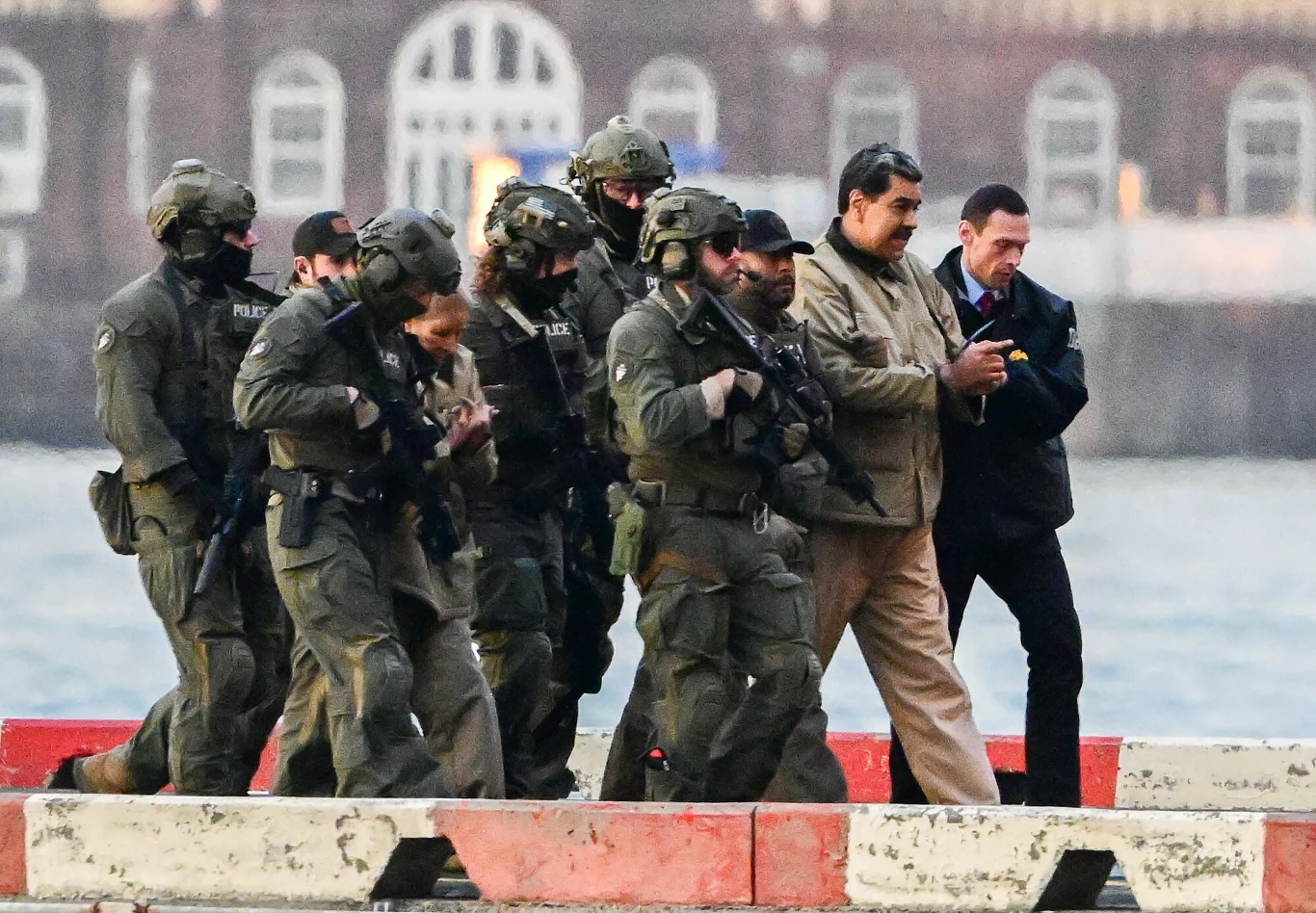 Nicolas Maduro w Nowym Jorku. Azjatyckie stolice protestują Oddział uzbrojonych funkcjonariuszy służb specjalnych eskortuje dwóch mężczyzn w cywilnych ubraniach, z których jeden jest zakuty w kajdanki. Tło stanowi zabudowa miejska oraz woda.