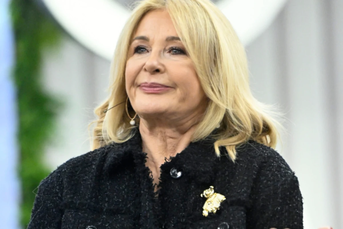 Majka Jeżowska Uśmiechnięta blondynka o dłuższych włosach ubrana w ciemną marynarkę z ozdobną broszką, stojąca w jasnym pomieszczeniu z rozmytym tłem.