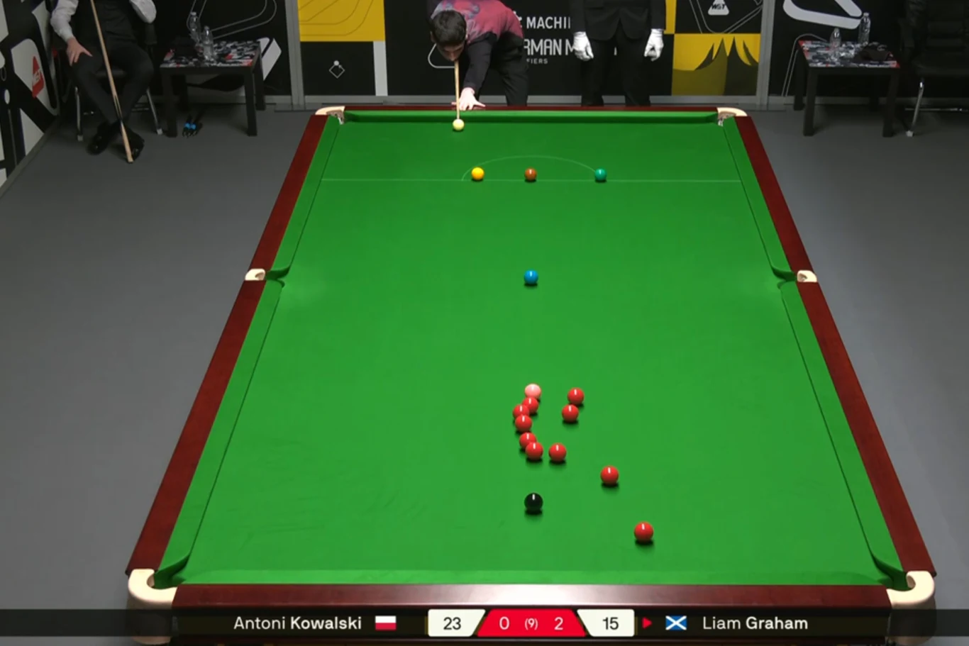 Zawodnik wykonuje uderzenie w trakcie meczu snookera, na stole rozmieszczone są bile o różnych kolorach, a w tle znajdują się sędziowie lub osoby towarzyszące. Wynik meczu i nazwiska zawodników widoczne na dole obrazu.