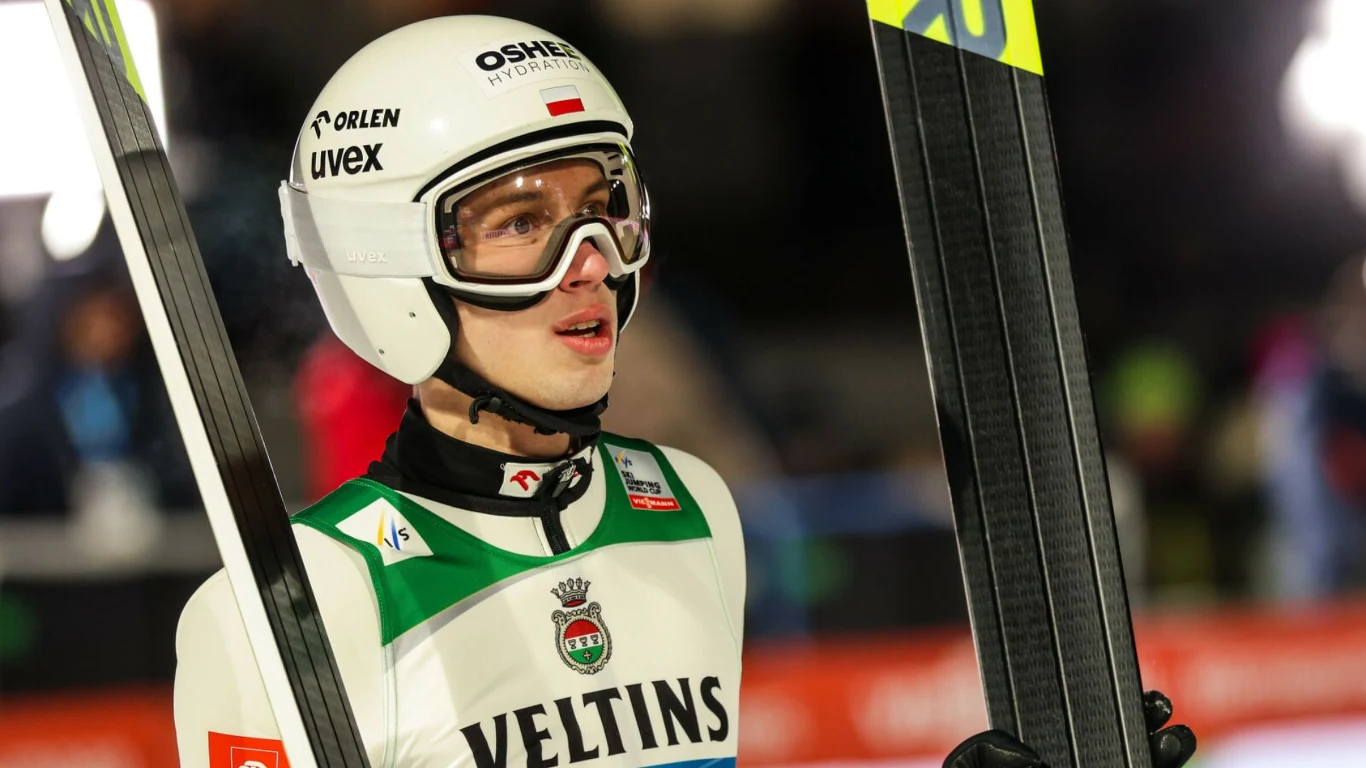 Kacper Tomasiak Skoczek narciarski w kombinezonie sportowym oraz kasku, trzymający narty, stoi na tle rozmytego stadionu z widocznymi światłami.