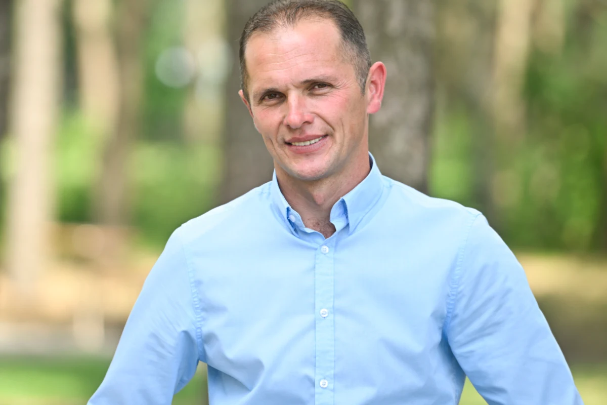 Waldemar z "Rolnik szuka żony" 10 Mężczyzna w jasnoniebieskiej koszuli stoi na tle rozmytego, zielonego lasu. Uśmiecha się i patrzy bezpośrednio w obiektyw, w jego postawie widać pewność siebie i spokój.