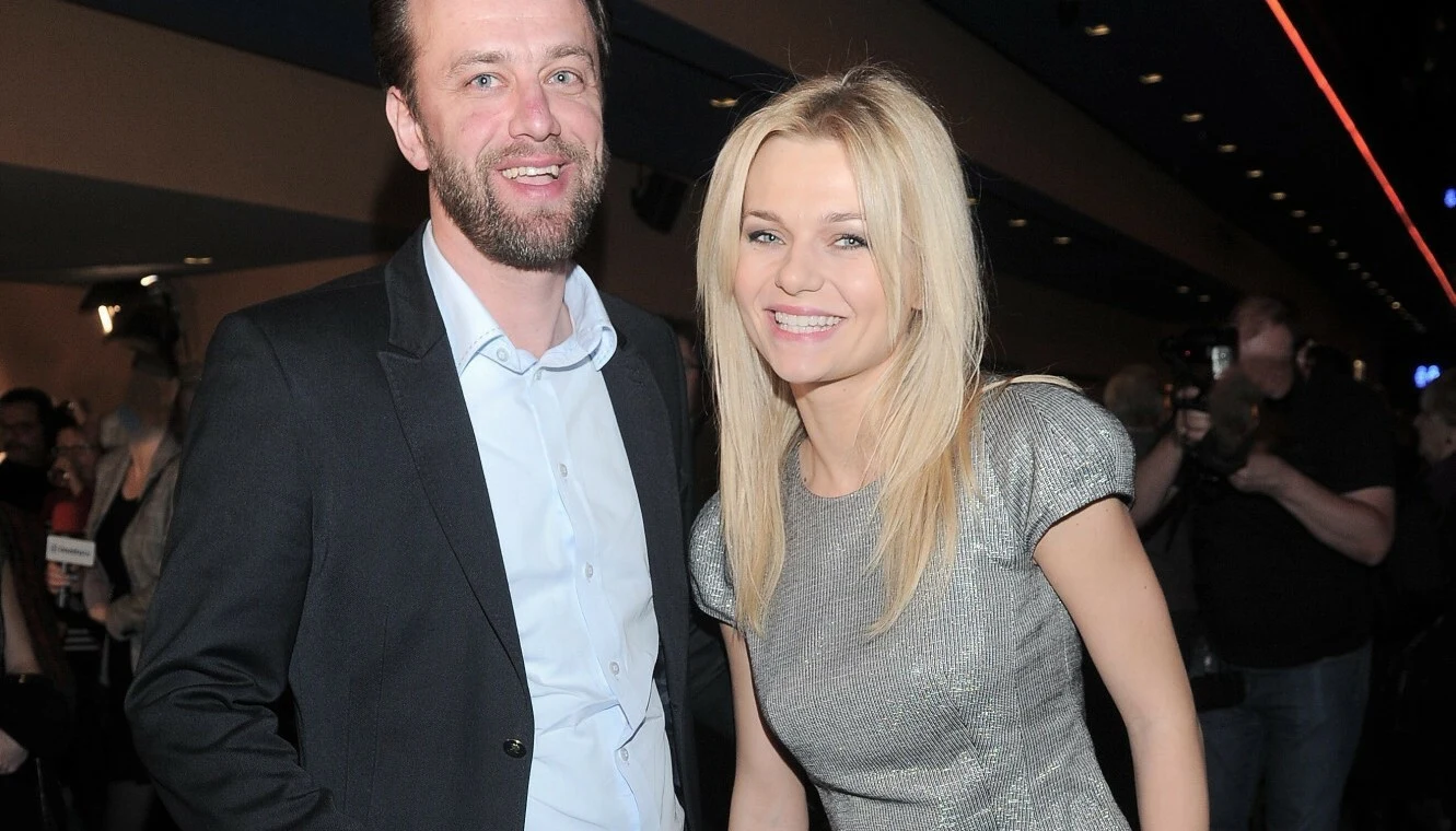 Łukasz Konopka i Emilia Komarnicka Uśmiechnięta kobieta w srebrnej sukience i mężczyzna w garniturze stoją razem w oświetlonym wnętrzu, w tle widoczna grupa ludzi i kamery.