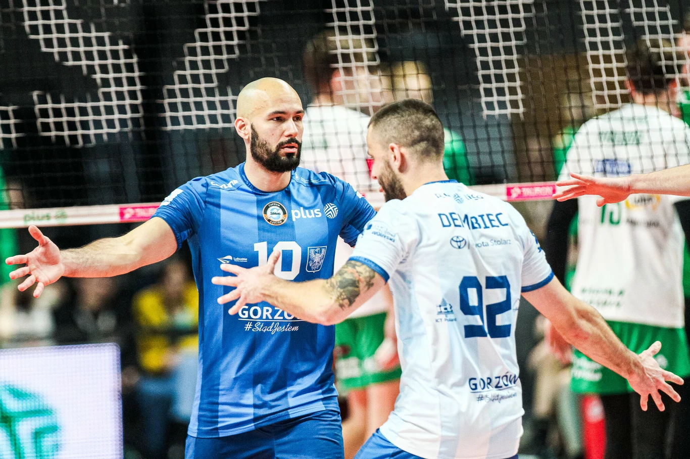 Siatkarze w sportowych strojach wykonują gest celebracyjny na boisku podczas meczu siatkówki, w tle siatka oraz inni zawodnicy.