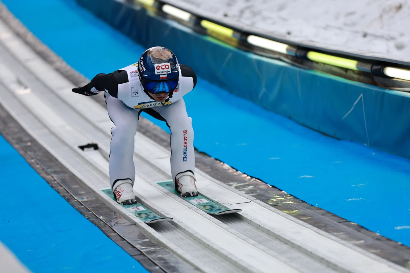 Zawodnik w kombinezonie narciarskim w pozycji najazdowej na rozbiegu skoczni narciarskiej, otoczony sztucznym podłożem oraz śniegiem, w tle widoczne bandy ochronne.