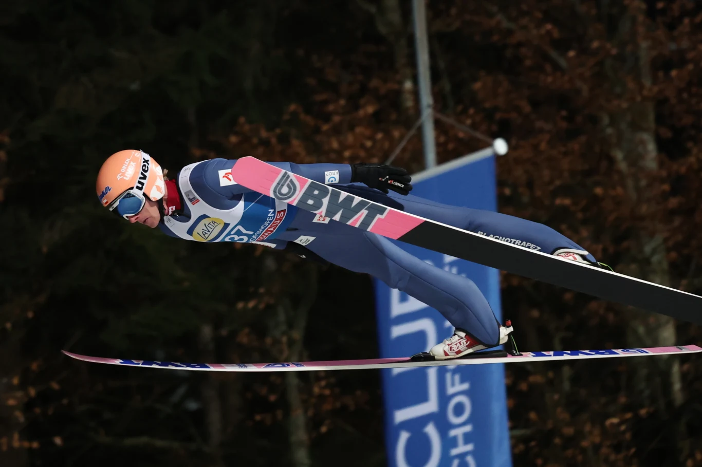 Zawodnik w niebieskim kombinezonie podczas lotu na nartach w trakcie skoku narciarskiego, w tle rozmazane drzewa i fragment baneru zawodów