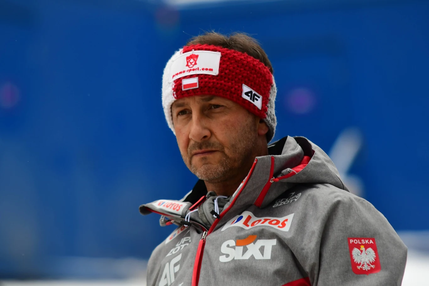 Mężczyzna w sportowej kurtce i czerwono-białej opasce z godłem Polski, stojący na tle niebieskiej ściany, uważny wyraz twarzy, liczne logotypy sponsorów na ubraniu.