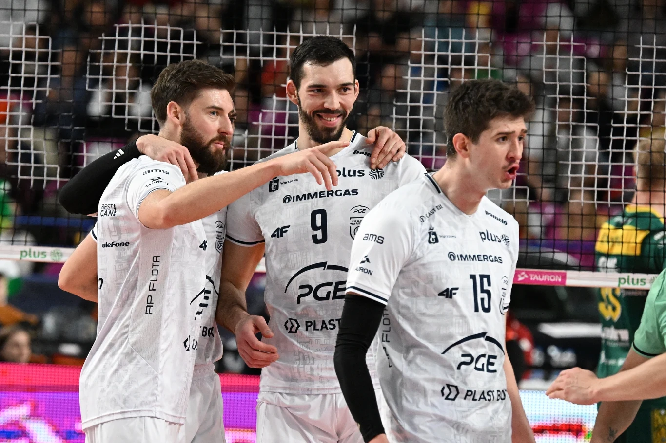 Trzech siatkarzy w białych strojach drużyny PGE Skra Bełchatów świętuje zdobycie punktu podczas meczu, a w tle widoczne są siatka oraz zawodnicy przeciwnej drużyny ubrani w zielone stroje.