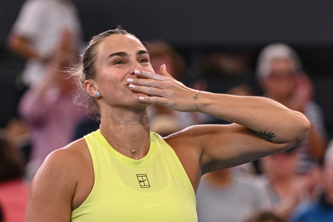 Kobieta w sportowym stroju na korcie tenisowym wykonuje gest pocałunku skierowanego do publiczności, w tle widoczny rozmyty tłum kibiców.