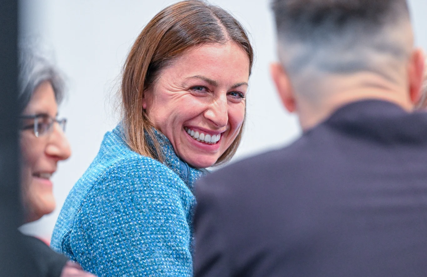 Justyna Kowalczyk-Tekieli Uśmiechnięta kobieta w niebieskim swetrze siedzi w towarzystwie dwóch osób, rozmawiając w przyjaznej atmosferze. W centrum kadru skupiona jest kobieta, która kieruje wzrok w stronę aparatu.