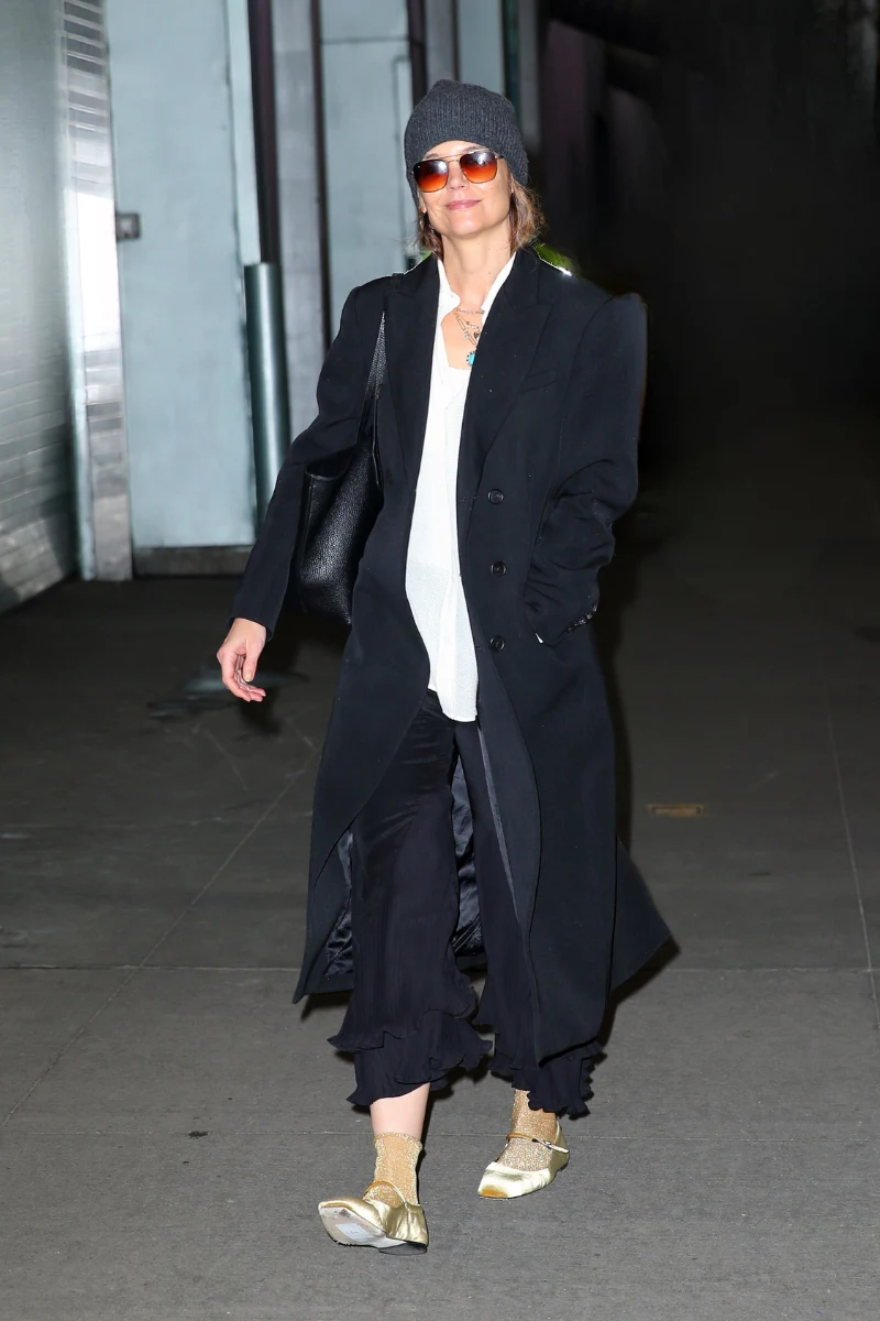 Katie Holmes w czarnym płaszczu, spodniach z falbankami, skarpetkach i złotych balerinach
