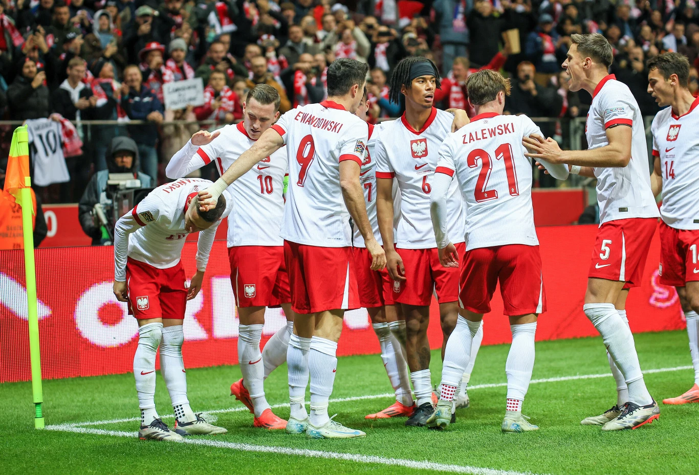 Maxi Oyedele (w środku) Grupa polskich piłkarzy w białych i czerwonych strojach świętuje zdobycie bramki na stadionie podczas meczu przy kibicujących tłumach w tle.