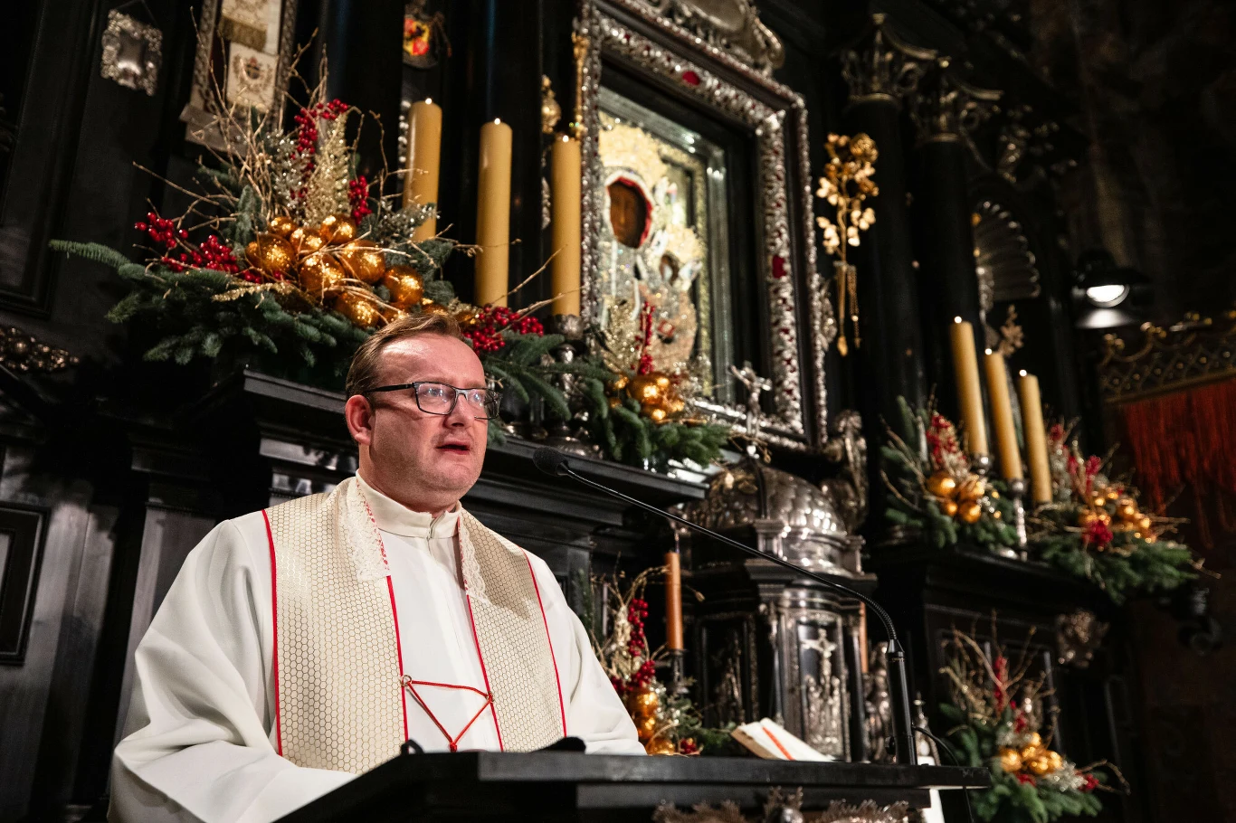 Kapłan ubran w białą albę i ornat stoi przy mównicy, za nim bogato zdobione ołtarze udekorowane złotymi świecami i świątecznymi stroikami z zielenią i czerwonymi akcentami, w tle obraz Matki Boskiej Częstochowskiej.