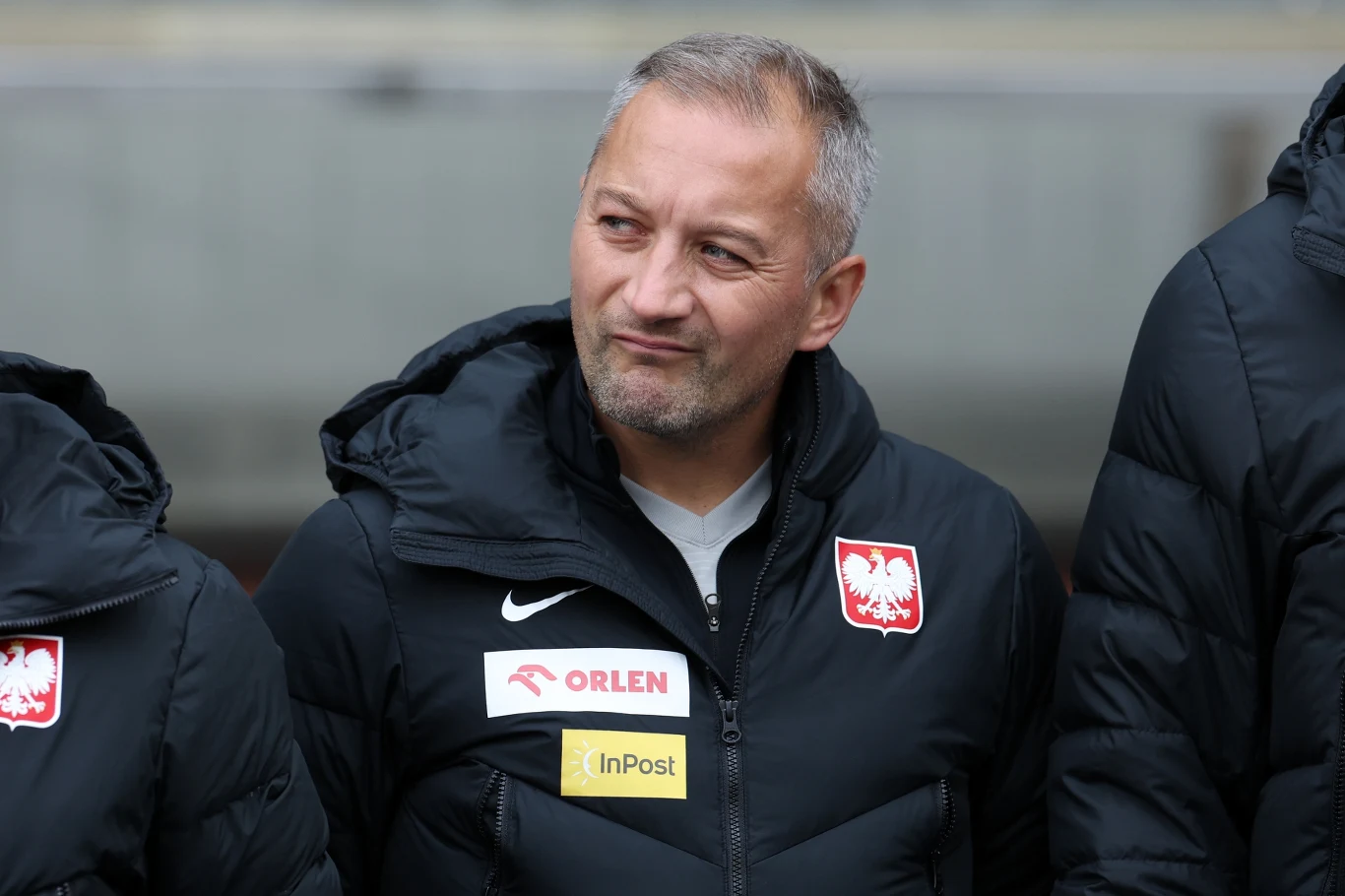Mężczyzna w ciemnej kurtce sportowej z godłem Polski oraz logotypami sponsorów Orlen i InPost, stojący obok innych osób w podobnych kurtkach, z lekkim uśmiechem i krótkimi siwymi włosami.