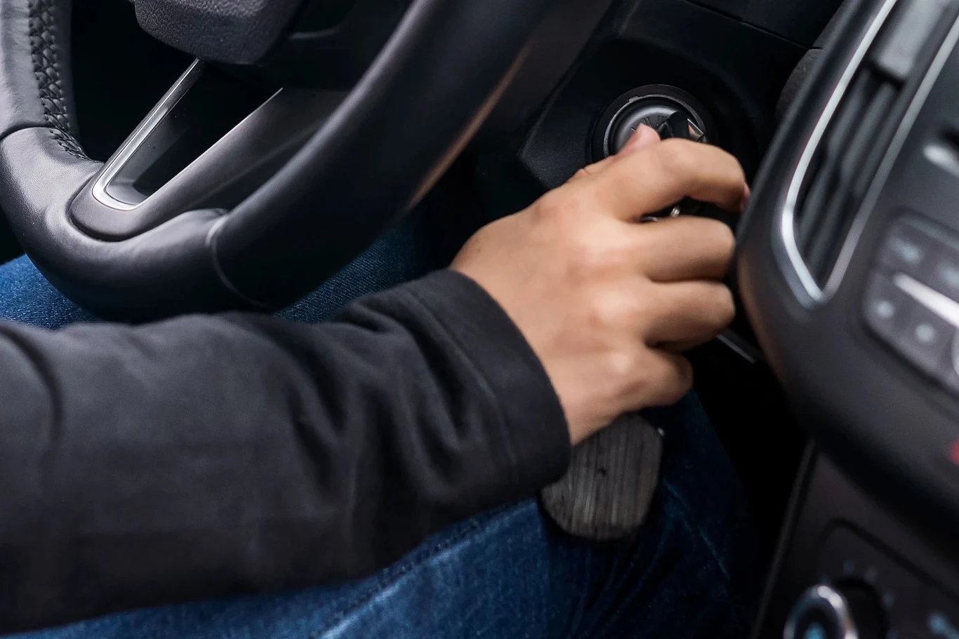 Zimą warto stosować metodę odpalania silnika na dwa razy Ręka w ciemnej bluzie przekręca kluczyk w stacyjce samochodu, kierownica i część deski rozdzielczej są widoczne na pierwszym planie.