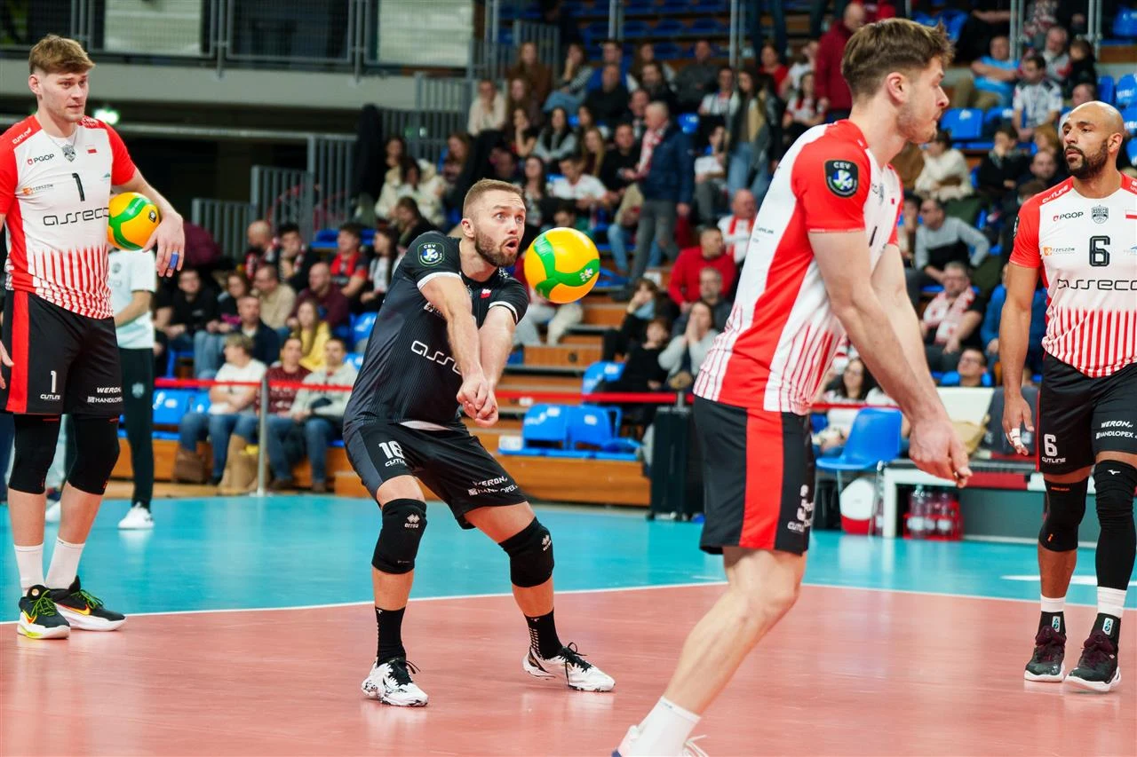 Zawodnicy drużyny siatkarskiej w trakcie meczu na hali sportowej, skupieni na przyjęciu piłki, w otoczeniu publiczności na trybunach. Atmosfera pełna emocji i sportowej rywalizacji.