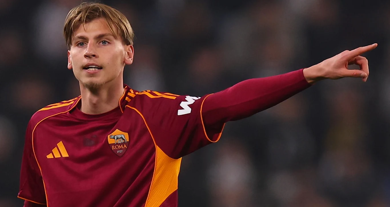 Jan Ziółkowski w barwach AS Roma Młody piłkarz w bordowej koszulce AS Roma z żółtymi detalami wskazuje ręką kierunek na boisku, koncentrując się na grze, w tle rozmyta publiczność stadionu.