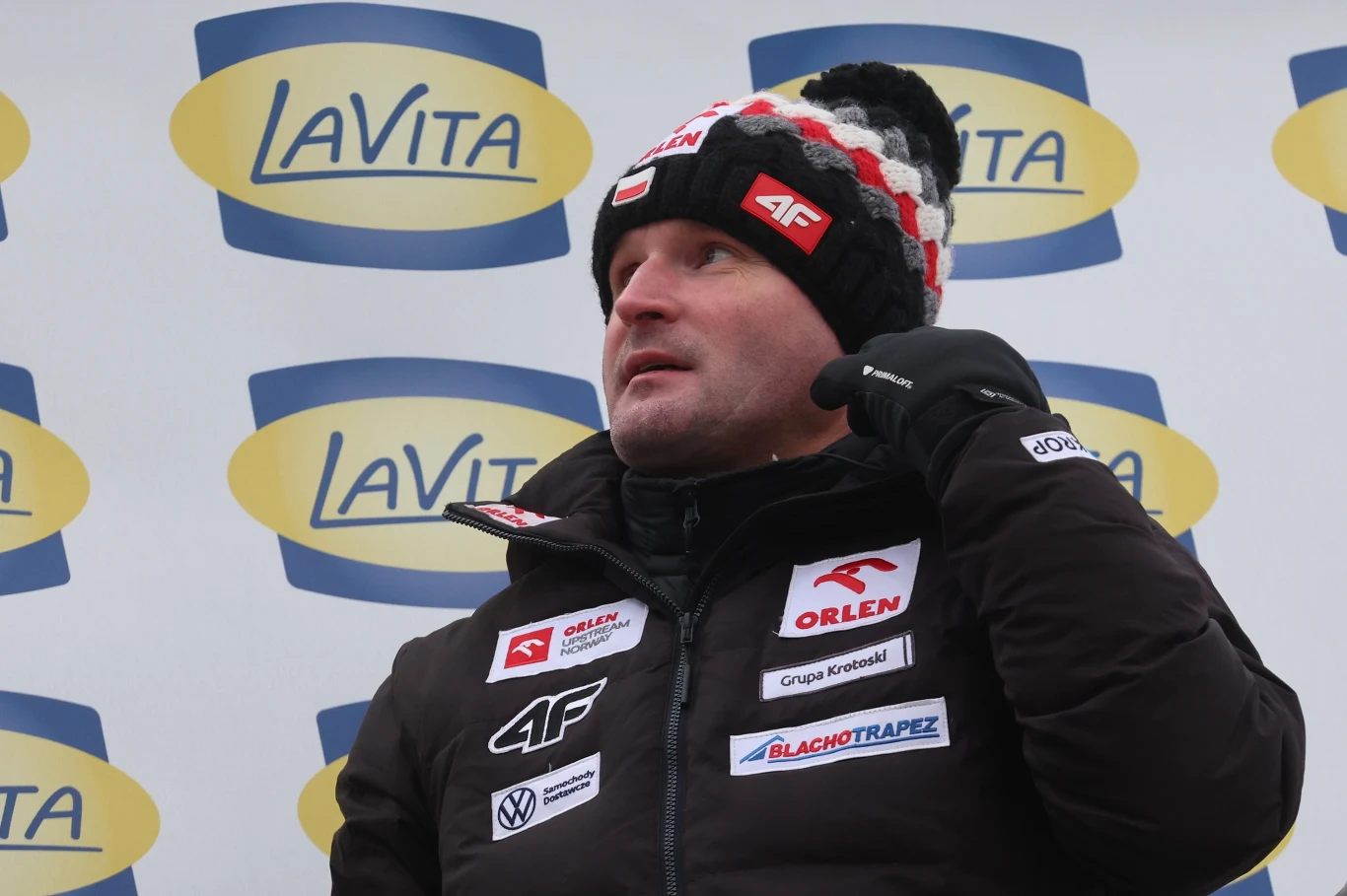 Mężczyzna w zimowej kurtce i czapce z polskimi akcentami stoi na tle ścianki z logo LaVita, jego kurtka pokryta jest naszywkami sponsorów sportowych.