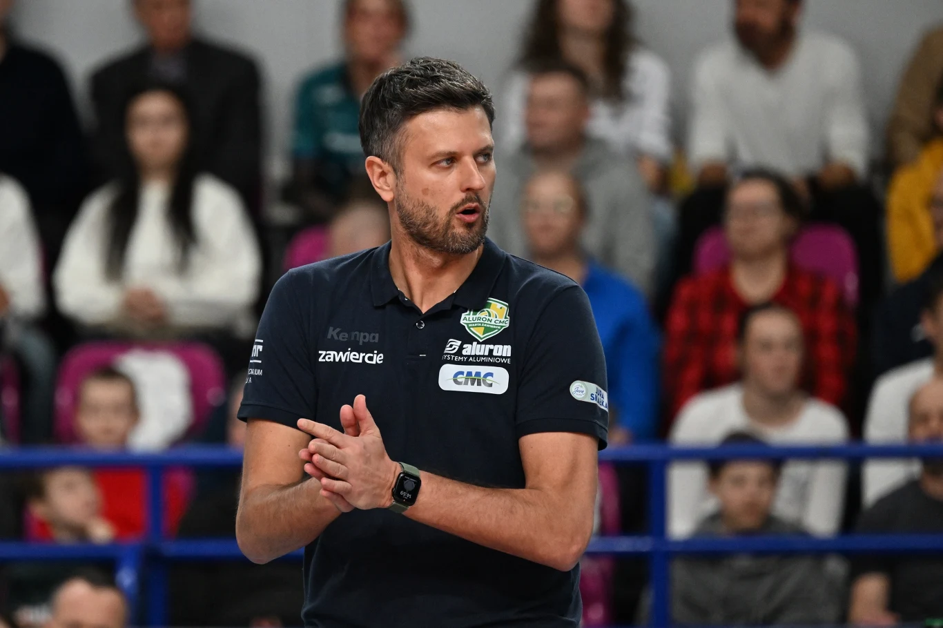 Michał Winiarski, trener Aluron CMC Warty Zawiercie Trener siatkarski w ciemnej koszulce z logami sponsorów stoi na hali sportowej, klaszcząc w dłonie, wokół widoczna publiczność siedząca na trybunach.