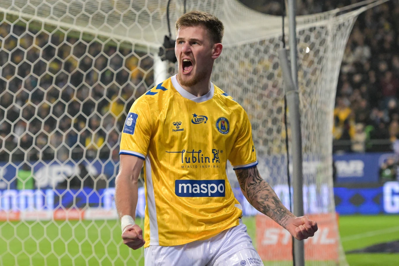 Piłkarz ubrany w żółtą koszulkę i białe spodenki świętuje zdobycie bramki na tle siatki bramkowej podczas meczu, w tle rozmazana publiczność.